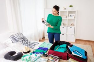 tourism, people and luggage concept - happy young woman packing travel bag at home or hotel room
